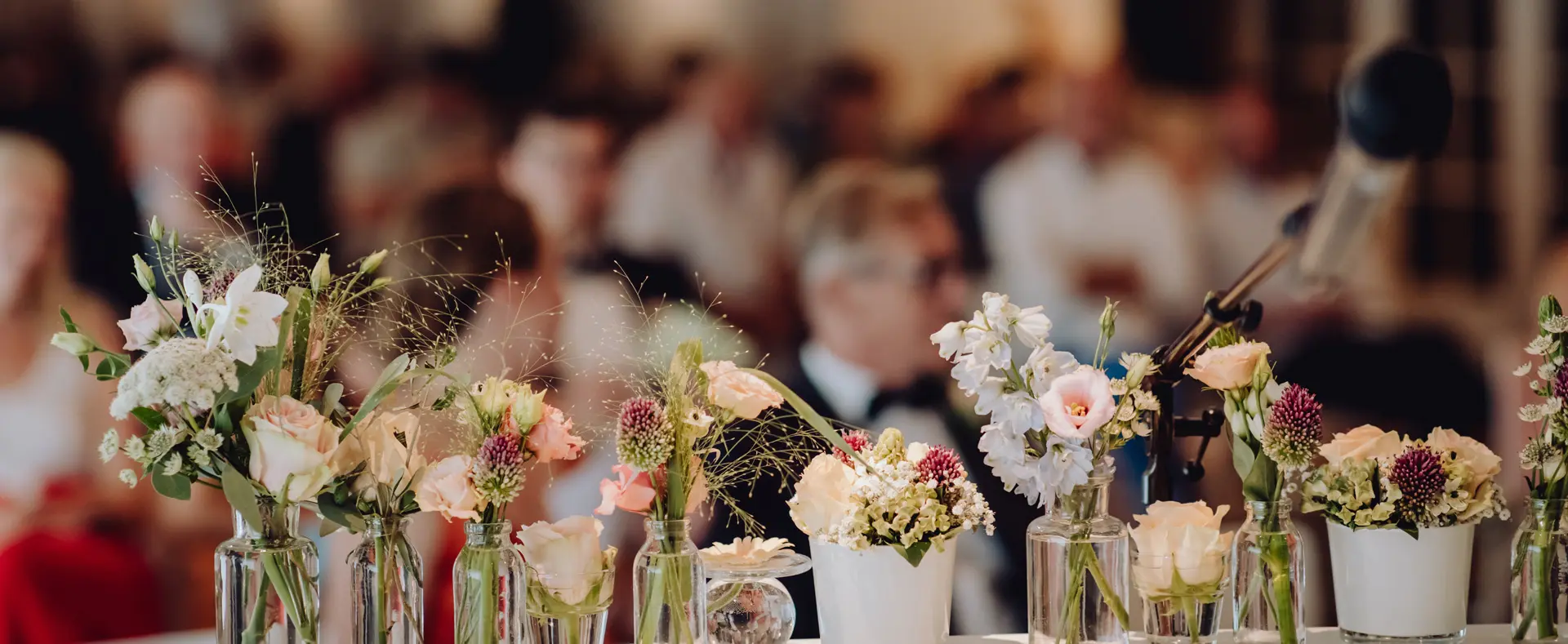 Bloemen in vaasjes op de ceremonietafel, met de gasten op de achtergrond